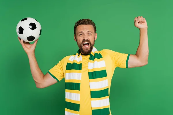 Emotionaler Fußballfan mit gestreiftem Schal hält Fußball in der Hand und jubelt isoliert auf Grün — Stockfoto