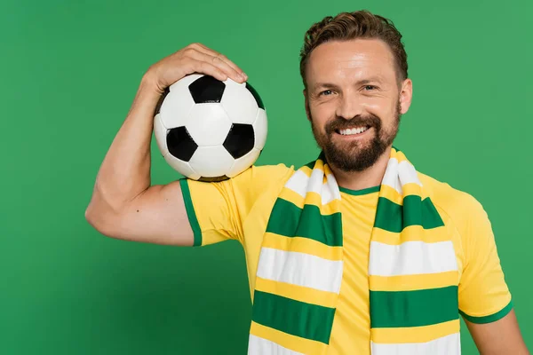 Улыбающийся футбольный фанат в полосатом шарфе и желтой футболке держа футбол изолирован на зеленый — стоковое фото