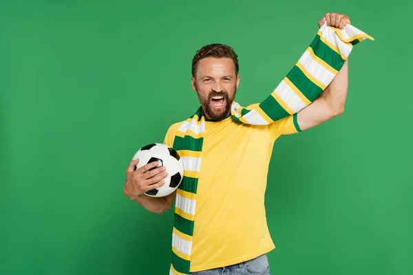 Схвильований футбольний вентилятор у смугастому шарфі, що тримає футбол ізольовано на зеленому — стокове фото