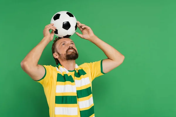 Bearded soccer fan in striped scarf and yellow t-shirt holding football on head isolated on green — Stock Photo