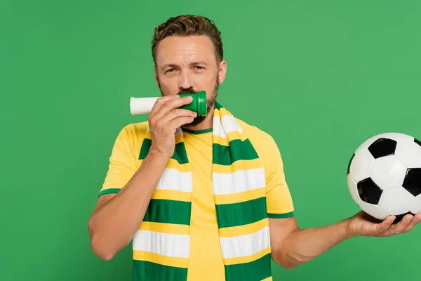 Bearded sports fan holding football and blowing horn while cheering isolated on green — Stock Photo