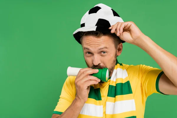 Barbudo hombre en fútbol sombrero soplando cuerno mientras mira la cámara aislado en verde - foto de stock