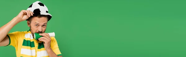 Barbudo hombre en el sombrero de fútbol soplando cuerno mientras anima aislado en verde, pancarta - foto de stock