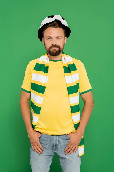 Homme barbu en chapeau de ventilateur de sport et écharpe rayée debout avec les mains dans des poches isolées sur vert — Photo de stock