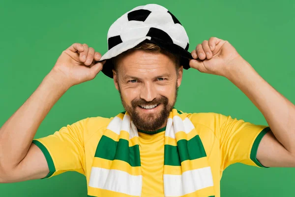 Homem barbudo feliz em cachecol listrado usando chapéu de fã de futebol isolado no verde — Fotografia de Stock
