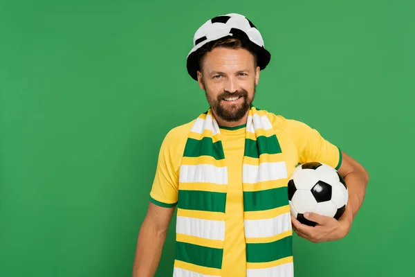 Homem barbudo feliz em chapéu de fã de futebol e cachecol listrado de pé com bola isolada no verde — Fotografia de Stock