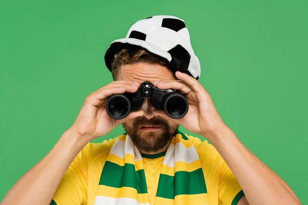 Bearded man in football fan hat and striped scarf looking through binoculars during match isolated on green — Stock Photo