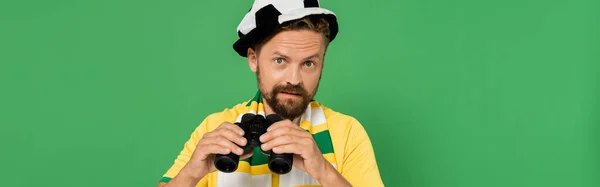Homme barbu en chapeau de ventilateur et écharpe rayée tenant des jumelles pendant le match de football isolé sur vert, bannière — Photo de stock