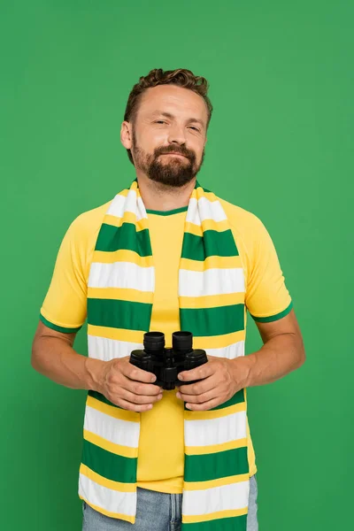 Hombre barbudo en bufanda a rayas sosteniendo binoculares durante el partido de fútbol aislado en verde - foto de stock