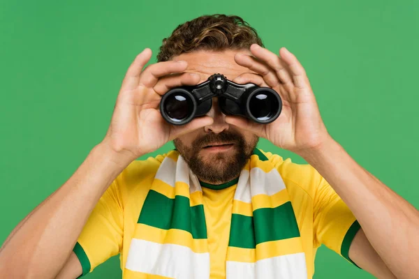 Homem barbudo em cachecol listrado olhando através de binóculos durante jogo de futebol isolado em verde — Fotografia de Stock