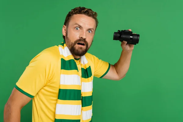 Удивленный мужчина в полосатом шарфе держит бинокль во время футбольного матча изолированы на зеленый — стоковое фото
