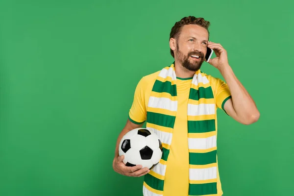 Hombre alegre en bufanda a rayas sosteniendo el fútbol y hablando en el teléfono móvil aislado en verde - foto de stock