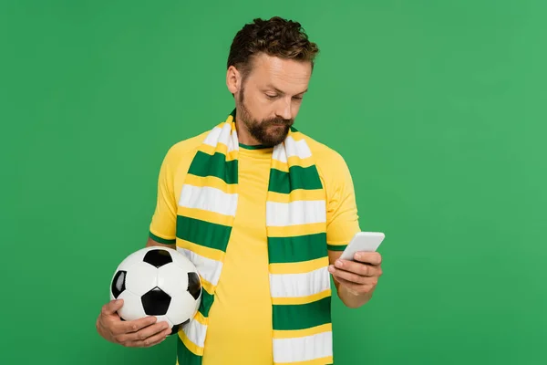 Bearded man in striped scarf holding football and using mobile phone isolated on green — Stock Photo