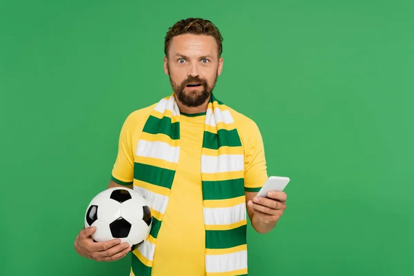 Ошеломленный мужчина в полосатом шарфе держа футбол и мобильный телефон изолированы на зеленый — стоковое фото
