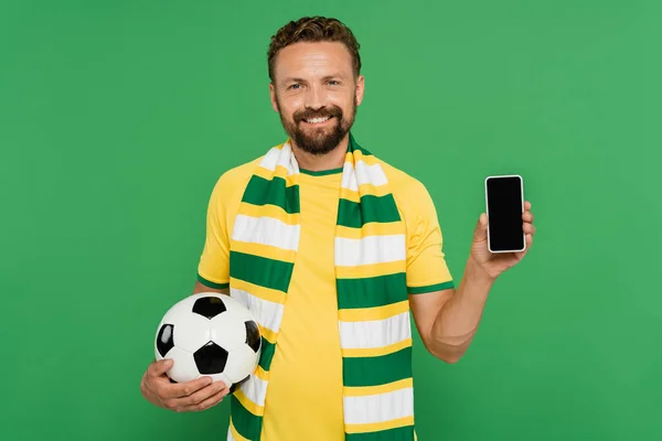 Веселий чоловік в смугастому шарфі тримає футбол і смартфон з порожнім екраном ізольовано на зеленому — стокове фото