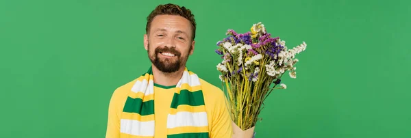 Alegre y barbudo ventilador de fútbol sosteniendo flores silvestres aislados en verde, bandera - foto de stock