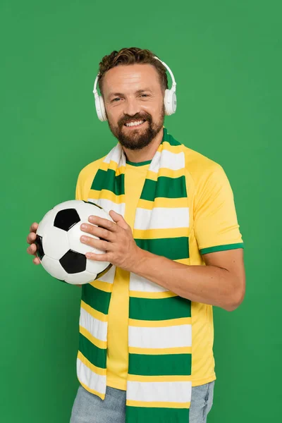 Щасливий бородатий спортивний вентилятор в шарфі і навушники, що тримають футбол ізольовано на зеленому — стокове фото