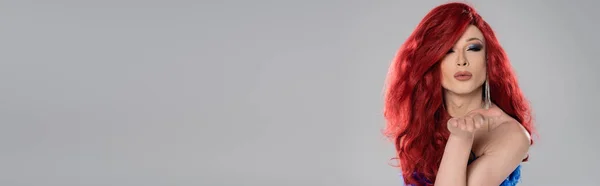 Drag queen en peluca roja soplando aire beso en la cámara aislado en gris, pancarta - foto de stock