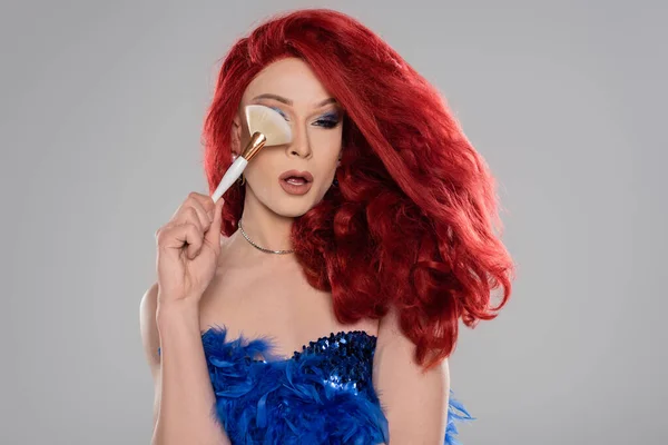 Drag queen élégant en perruque tenant brosse de maquillage près du visage isolé sur gris — Photo de stock