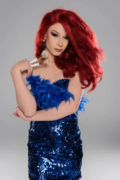Элегантная трансвестит в парике и синем платье с кисточками для макияжа, изолированными на сером — стоковое фото