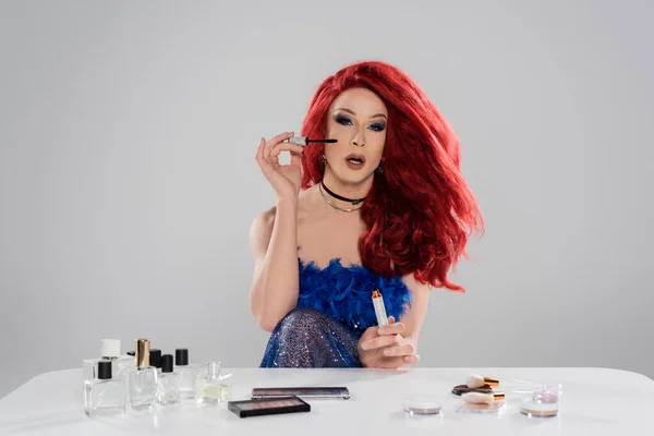 Drag queen in red wig holding mascara near decorative cosmetics and perfumes isolated on grey — Stock Photo