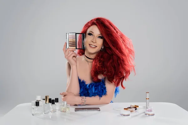 Улыбающаяся королева-трансвестит держит тени для век рядом с косметикой и парфюмерией, изолированными на сером — стоковое фото