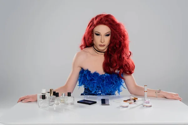 Элегантный трансвестит в красном парике, смотрящий на камеру рядом с декоративной косметикой и парфюмерией, изолированной на сером — стоковое фото