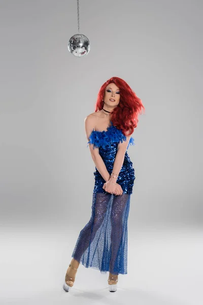 Улыбающаяся трансвестит в платье и на каблуках смотрит в камеру возле диско-мяча на сером фоне — стоковое фото