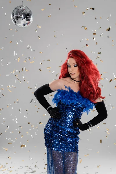 Smiling drag queen in elegant dress and gloves posing under confetti and disco ball on grey background — Stock Photo