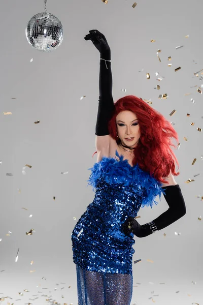 Cheerful drag queen in gloves and dress posing under confetti and disco ball on grey background — Stock Photo