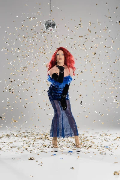 Элегантный трансвестит в платье и перчатках, дующий воздушный поцелуй под диско мяч и конфетти на сером фоне — стоковое фото