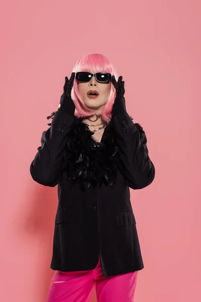 Drag queen en peluca y chaqueta tocando gafas de sol sobre fondo rosa - foto de stock