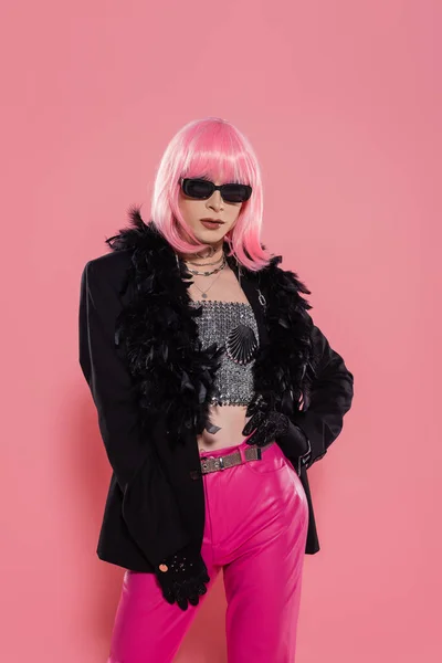 Trendy drag queen in sunglasses and jacket with feathers standing on pink background — Stock Photo