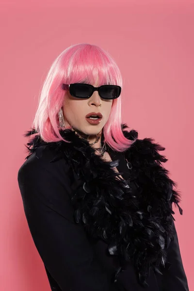 Retrato de persona transgénero con estilo con maquillaje y gafas de sol posando en chaqueta con plumas sobre fondo rosa - foto de stock