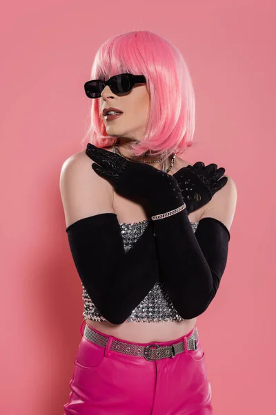 Retrato de drag queen de moda en gafas de sol y guantes tocando hombros sobre fondo rosa - foto de stock