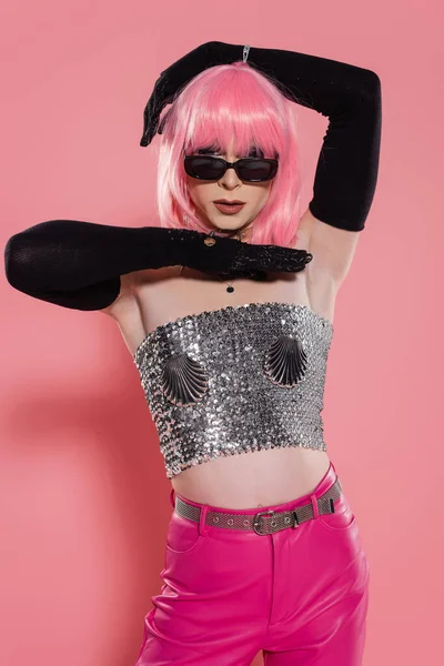Retrato de drag queen en gafas de sol y top plateado brillante posando sobre fondo rosa - foto de stock
