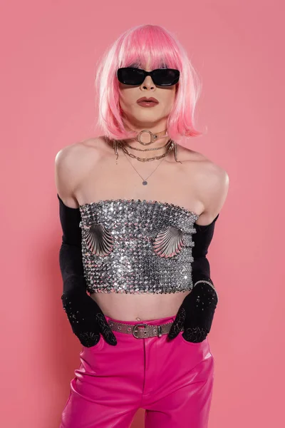Portrait of stylish drag queen in silver top and gloves holding hands in pockets of pants on pink background — Stock Photo