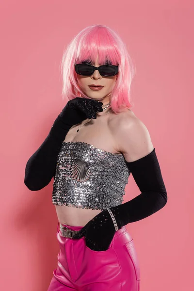 Retrato de drag queen de moda en la parte superior brillante y guantes tocando la barbilla sobre fondo rosa - foto de stock
