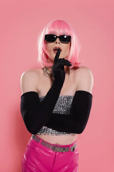 Drag queen élégant dans des lunettes de soleil et des gants montrant un geste de silence sur fond rose — Photo de stock
