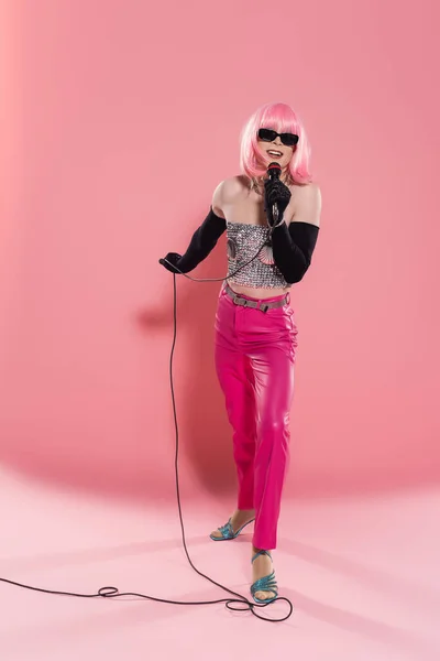 Longitud completa de drag queen sonriente en guantes y gafas de sol con micrófono sobre fondo rosa - foto de stock