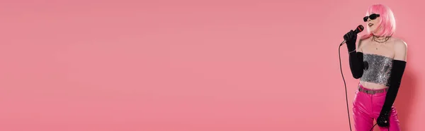 Стильный трансвестит в солнцезащитных очках и перчатках держа микрофон на розовом фоне, баннер — стоковое фото