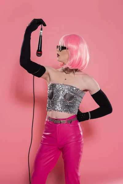 Vista lateral de drag queen na moda em óculos de sol segurando microfone no fundo rosa — Fotografia de Stock
