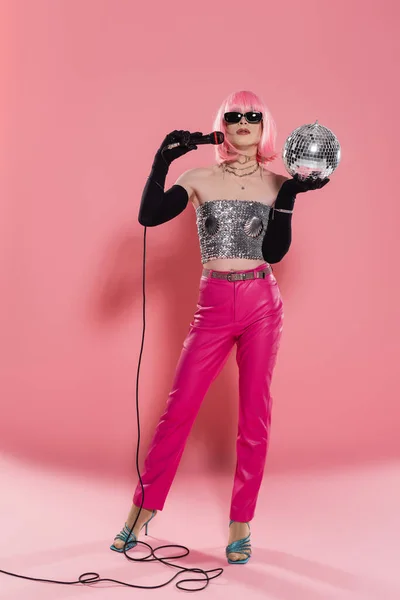 Full length of trendy drag queen in sunglasses and gloves holding microphone and disco ball on pink background — Stock Photo