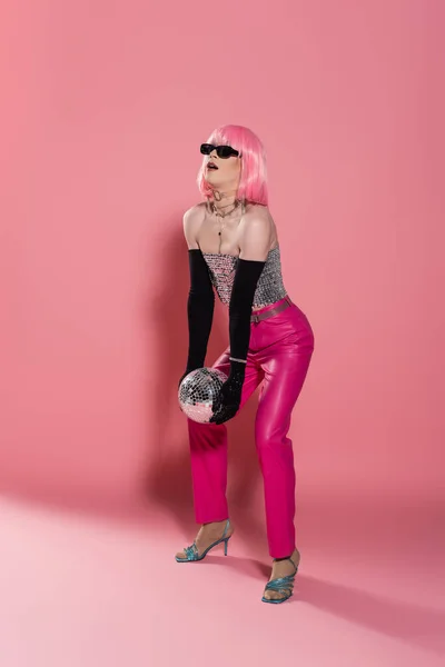 Fashionable drag queen in wig and sunglasses holding heavy disco ball on pink background — Stock Photo