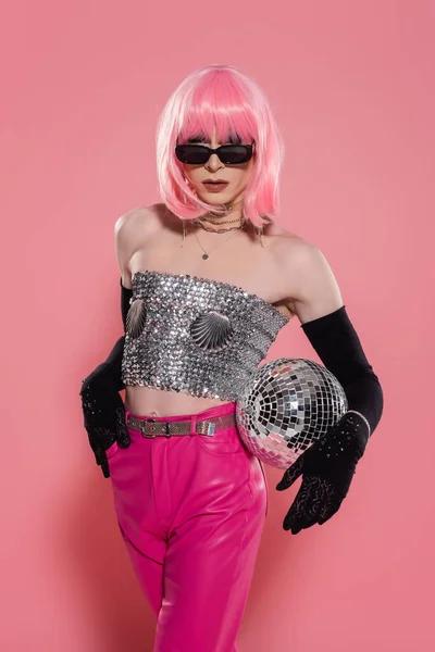 Модная королева драга в солнцезащитных очках и перчатках, держащая дискотечный мяч на розовом фоне — стоковое фото