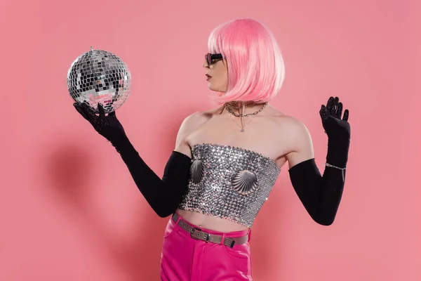 Vista lateral de la drag queen de moda en la parte superior brillante y guantes que sostienen la bola disco sobre fondo rosa - foto de stock