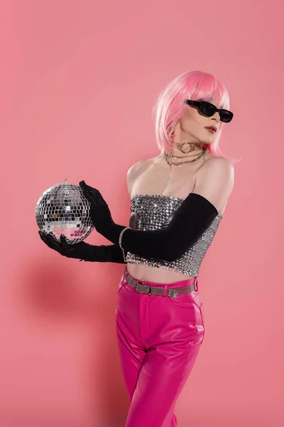 Fashionable drag queen in sunglasses and silver top holding disco ball on pink background — Stock Photo
