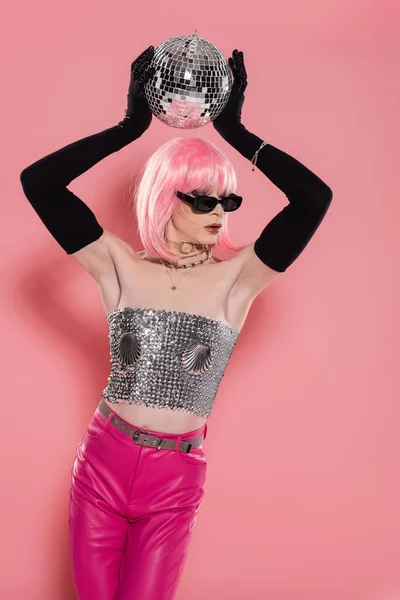 Хорошо одет трансвестит в перчатках и солнцезащитных очках держа диско мяч на розовом фоне — стоковое фото