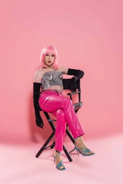 Fashionable drag queen in shiny top sitting on chair on pink background — Stock Photo
