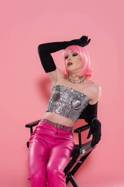 Fashionable drag queen in top and gloves posing while sitting on chair on pink background — Stock Photo